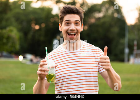 Immagine di uomo felice 20s a piedi attraverso il parco e bere una bevanda dalla tazza di plastica Foto Stock