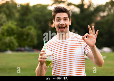 Immagine del giovane uomo 20s a piedi attraverso il parco e bere una bevanda dalla tazza di plastica Foto Stock