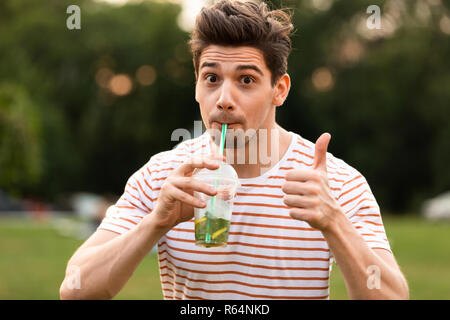 Immagine dell uomo divertente 20s a piedi attraverso il parco e bere una bevanda dalla tazza di plastica Foto Stock