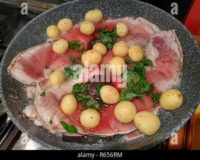 In padella le fette di pesce spada, condito con prezzemolo fresco in foglie, le patate di primizia, pepe. La cucina si muove le fette, un po' di vapore sorge dal Foto Stock