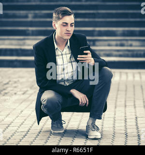 Bel giovane uomo d affari utilizzando smart phone seduto in strada della citta'. Alla moda ed elegante modello maschile in giacca nera Foto Stock