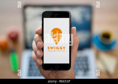 Un uomo guarda al suo iPhone che visualizza il logo Swiggy, mentre se ne sta seduto alla sua scrivania per computer (solo uso editoriale). Foto Stock