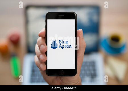 Un uomo guarda al suo iPhone che visualizza il grembiule blu logo, mentre se ne sta seduto alla sua scrivania per computer (solo uso editoriale). Foto Stock