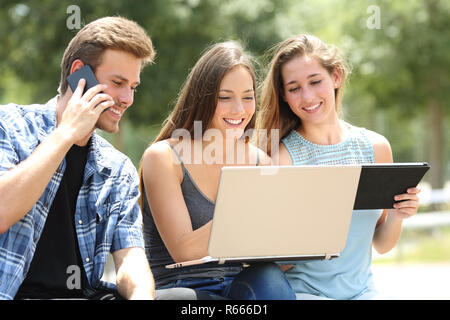 Tre happy amici utilizzando più dispositivi seduta su una panchina nel parco Foto Stock