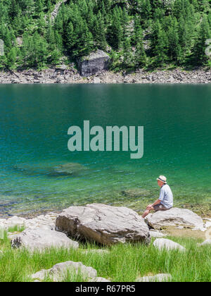 Uomo anziano, indossando un cappello di Panama, seduto da solo su una roccia da un lago. Foto Stock