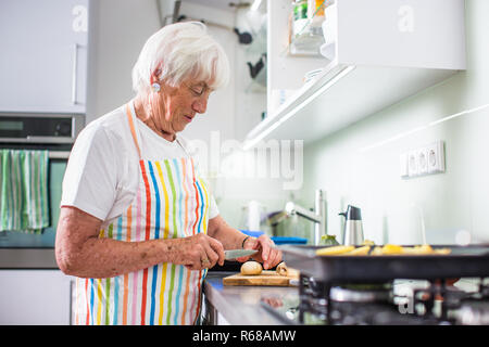 Senior donna cucina in cucina - mangiare e cucinare sano per la sua famiglia mettendo alcune potates nel forno, godendo di pensionamento attivo (SHALLOW DOF dai toni di colore immagine) Foto Stock