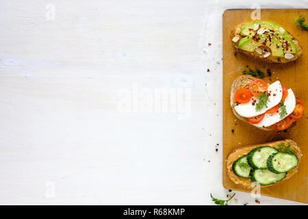 Toast freschi panini con cetrioli, avocado e pomodori in pezzi di forma ovale di pane scuro, visto dal di sopra con il bianco la superficie del tavolo per spazio di copia Foto Stock