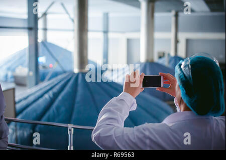 Donna in tuta. Un dipendente in una veste bianca prende una foto negozio in fabbrica sul territorio dell'impianto di produzione industriale, acciaio serbatoi di fermentazione Foto Stock