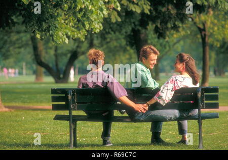 Ritratto, Parkszene, junge Frau mit langen Haaren braunen flirtet mit 2 jungen Maennern auf einer Parkbank Foto Stock