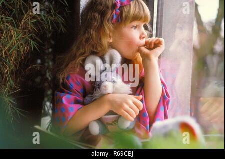 Ritratto, Maedchen mit langen braunen Haare, 7 Jahre, sitzt traurig mit ihrem Stofftier daumenlutschend am Fenster Foto Stock