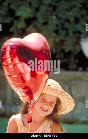 Ritratto, all'aperto, Maedchen mit langen Haaren braunen, 6 Jahre alt, mit Strohhut haelt einen herzfoermigen roten Luftballon un einer Schnur fest Foto Stock