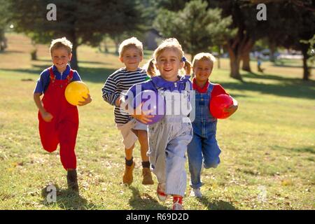 All'aperto, Parkszene, Gruppe von vier Kindern, 5-8 Jahre, laeuft mit bunten Luftballons unter dem braccio ueber eine Wiese Foto Stock