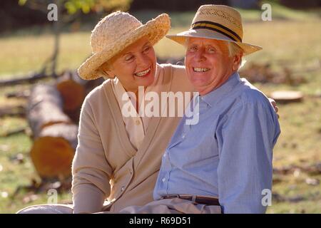 Ritratto, Halbfigur, Seniorenpaar bekleidet mit heller Kleidung und Stohhueten sitzt gluecklich zusammen auf einer Lichtung im Wald Foto Stock