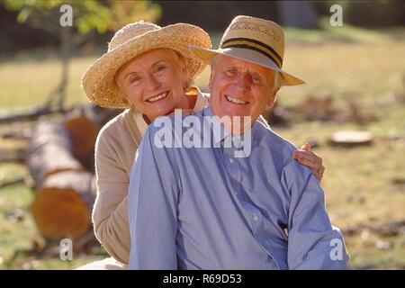 Ritratto, Halbfigur, Seniorenpaar bekleidet mit heller Kleidung und Stohhueten sitzt gluecklich zusammen auf einer Lichtung im Wald Foto Stock