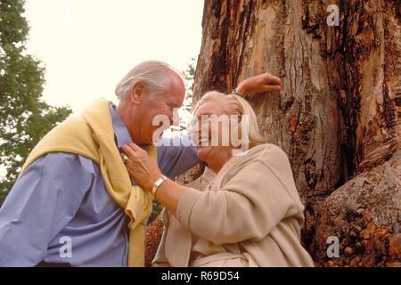 Ritratto, Halbfigur, Seniorenpaar bekleidet mit heller Kleidung unterhaelt sich glueckliches an den Stamm eines dicken Baumes angelehnt Foto Stock