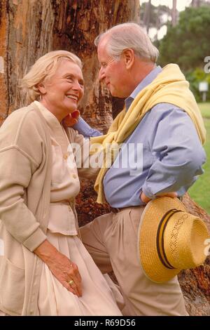 Ritratto, Halbfigur, Seniorenpaar bekleidet mit heller Kleidung unterhaelt sich glueckliches an den Stamm eines dicken Baumes angelehnt Foto Stock