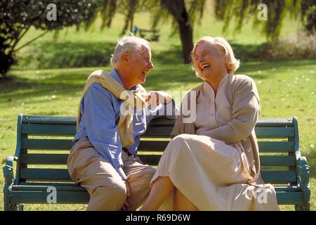 Parkszene, Outdoor, Ritratto, Halbfigur, Seniorenpaar bekleidet mit heller Kleidung unterhaelt sich lachend auf einer Parkbank Foto Stock