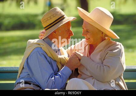 Parkszene, Outdoor, Ritratto, Halbfigur, Seniorenpaar bekleidet mit heller Kleidung und Strohhueten schmust zaertlich auf einer Parkbank Foto Stock