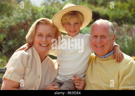 All'aperto, Ritratto, 6 Jahre alter blondr Junge mit Strohhut zwischen seinen auf einer Parkbank sitzenden Grosseltern Foto Stock