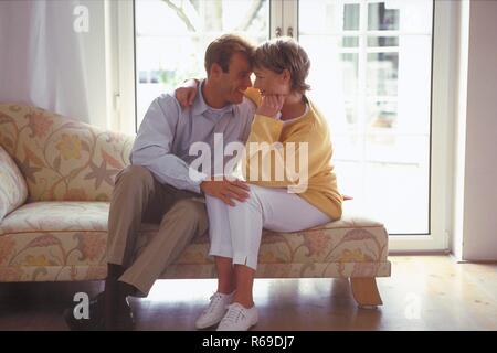 Ritratto, Innenraum, Ganzfigur, Profil, sich zaertlich anschauendes Paar Mitte 30 sitzt auf dem divano vor einer Glastuer Foto Stock
