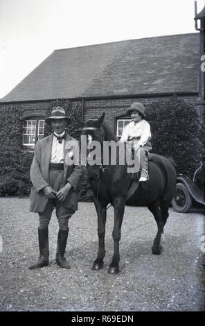 1930s, storico su una stradina di ghiaia al di fuori di una casa di campagna, un uomo anziano in marcia di equitazione in piedi accanto a una bambina su un pony o cavallo di piccole dimensioni, anche indossare abbigliamento da equitazione, probabilmente il suo nonno aiutarla a imparare a cavalcare. Foto Stock