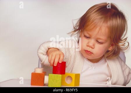 Ritratto, Innenraum, Halbfigur, bionde 2-jaehriges weiss gekleidetes Maedchen sitzt im Kinderstuhl und spielt mit bunten Holzkloetzen Foto Stock