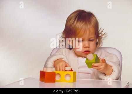 Ritratto, Innenraum, Halbfigur, bionde 2-jaehriges weiss gekleidetes Maedchen sitzt im Kinderstuhl und spielt mit bunten Holzkloetzen Foto Stock