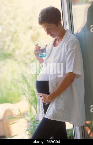 Ritratto, Halbfigur, schwangere Frau mit kurzen braunen Haaren bekleidet mit weissem Hemd und schwarzer Leggings steht Mit einem Glas Wasser an der Tuer zum Garten Foto Stock