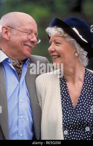 Ritratto, all'aperto, Profil, sich zaertlich anschauendes ca 75 Jahre altes Seniorenpaar, Mann mit Schnauzbart und Brille bekleidet mit Hemd gestreiftem, giacca und Halstuch und weisshaarige Frau im blauen Kleid, heller Jacke und blauem Hut Foto Stock