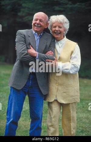Ritratto, Halbfigur, Outdoor, ca 75 Jahre altes Seniorenpaar im Park, Mann mit Schnauzbart bekleidet mit Jeans und camicia und weisshaarige Frau in gelber Hosenkombination Foto Stock