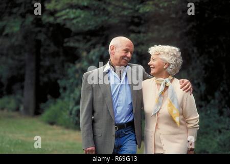 Ritratto, Halbfigur, Outdoor, ca 75 Jahre altes Seniorenpaar geht a braccetto im Park spazieren, Mann mit Schnauzbart bekleidet mit Jeans und camicia und weisshaarige Frau in gelber Kombination Foto Stock