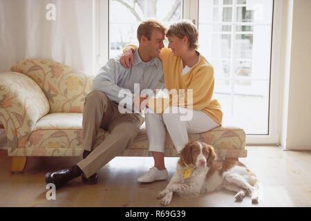 Ritratto, Innenraum, Ganzfigur, Profil, sich zaertlich anschauendes Paar Mitte 30 sitzt auf dem divano vor einer Glastuer, ihr Jagdhund liegt davor Foto Stock