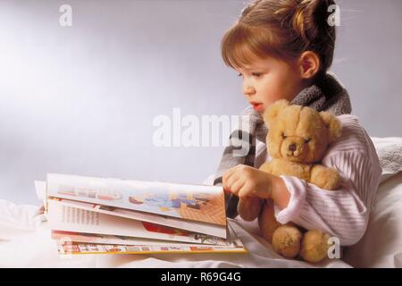 Ritratto, Innenraum, Profil, 7-jaehriges Maedchen bekleidet mit weissem Schlafanzug sitzt mit ihrem Teddy braccio im Bett im und liest ein Bilderbuch Foto Stock