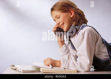 Ritratto, Innenraum, Profil, 12-jaehriges bionde Maedchen bekleidet mit weissem Shirt und Jeanslatzhose telefoniert Mit einem Handy waehrend sie ihre Hausaufgabe macht Foto Stock