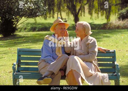 Parkszene, Outdoor, Ritratto, Halbfigur, Seniorenpaar bekleidet mit heller Kleidung unterhaelt sich lachend auf einer Parkbank, er traegt Strohhut Foto Stock