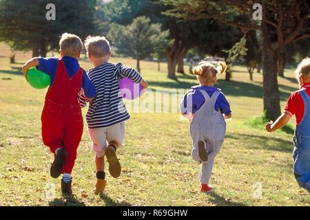 All'aperto, Parkszene, Gruppe von vier Kindern, 5-8 Jahre, laeuft mit bunten Luftballons unter dem braccio ueber eine Wiese von hinten gesehen Foto Stock