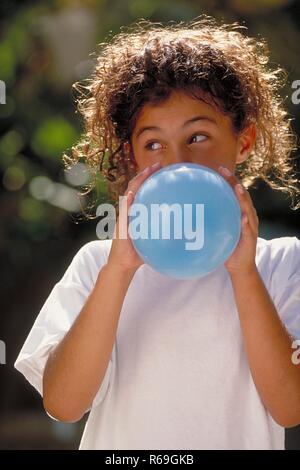 Ritratto, all'aperto, Maedchen mit hochgesteckten langen braunen Locken, 8 Jahre alt, bekleidet mit weissem T-Shirt blaest einen blauen Luftballon auf Foto Stock