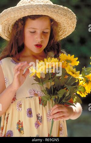 Ritratto, all'aperto, Halbfigur, Maedchen mit langen Haaren braunen, 6 Jahre alt, bekleidet mit Sommerkleid und Strohhut Mit einem Strauss gelber Blumen Foto Stock
