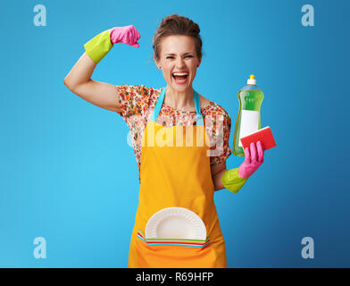 Felice casalinga nel grembiule arancione con spugna e detersivo per i piatti che mostra bicipite su sfondo blu. Casalinga soddisfatto del risultato degli strumenti selezionati - dishwas Foto Stock