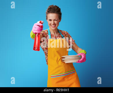 Sorridente giovane donna in grembiule arancione con piastre lavate utilizzando detergente per stoviglie come una pistola isolato sul blu. Donna felice con il nuovo altamente efficace di Foto Stock