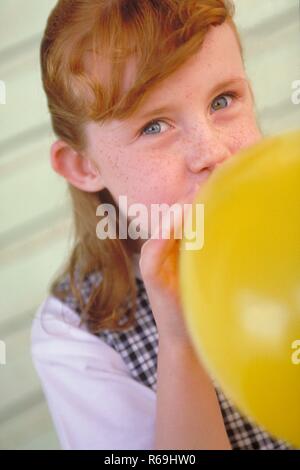 Ritratto, Nahaufnahme, sommersprossiges Maedchen mit langem roten Haar und blauen Augen, 6 Jahre, bekleidet mit weissem Hemd und blau-weiss kariertem Kleid sitzt vor einer gruengrau gestrichenen Bretterfassade und blaest einen gelben Luftballon auf Foto Stock