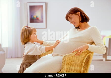 Maedchen, 3 Jahre, fuehlt die Bewegungen des Bambinos im Bauch circuizione hochschwangeren Mutter, die im Wohnzimmer in einem Korbsessel sitzt Foto Stock