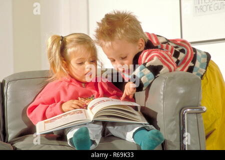 Maedchen, 2,5 Jahre sitzt Mit einem Bilderbuch auf den Knien auf dem divano, ihr Bruder, 4 Jahre, beugt sich ueber die Lehne und zeigt mit dem Finger auf ein Bild Foto Stock