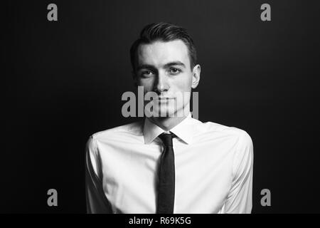 Chiudere fino in bianco e nero ritratto di un giovane uomo in camicia bianca e cravatta nera, seriamente guardando la telecamera, contro la pianura di sfondo per studio Foto Stock