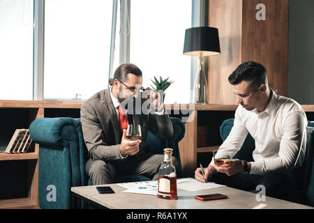 Due partner commerciali di sedere a tavola in ufficio e bere cognac Foto Stock