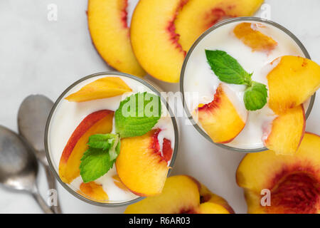 Due bicchieri di sano yogurt alla frutta con pesche fresche, cucchiai e menta su marmo bianco tavola. Colazione sana. vista superiore Foto Stock