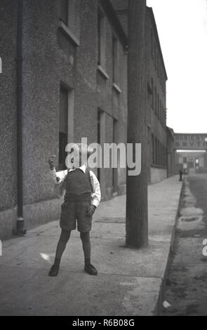 1940s, storico, eccitato giovane ragazzo in pantaloncini corti a giocare al di fuori sul marciapiede in strada urbana di case a schiera e il deposito, con un segno per i fabbricanti di maglieria Smyth & Co in distanza, una delle più antiche aziende di calzetteria in tutto il mondo e i fornitori del Queen's calze, Foto Stock