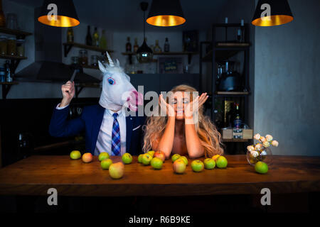 Coppia divertente giocare al bancone bar in cucina con le mele sulla tavola. Ragazza bionda divertirsi con il mio ragazzo in maschera. Unicorn in tuta con giovane donna Foto Stock