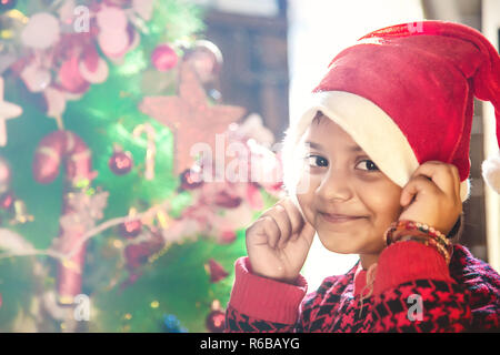 Little Indian/Asia kid ragazza in santa hat con albero di Natale e le luci sul background. Foto Stock