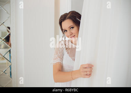 Bella bruna sposa in seta vestaglia e lacy velo holding abito da sposa su un appendiabiti vicino alla finestra. Foto Stock